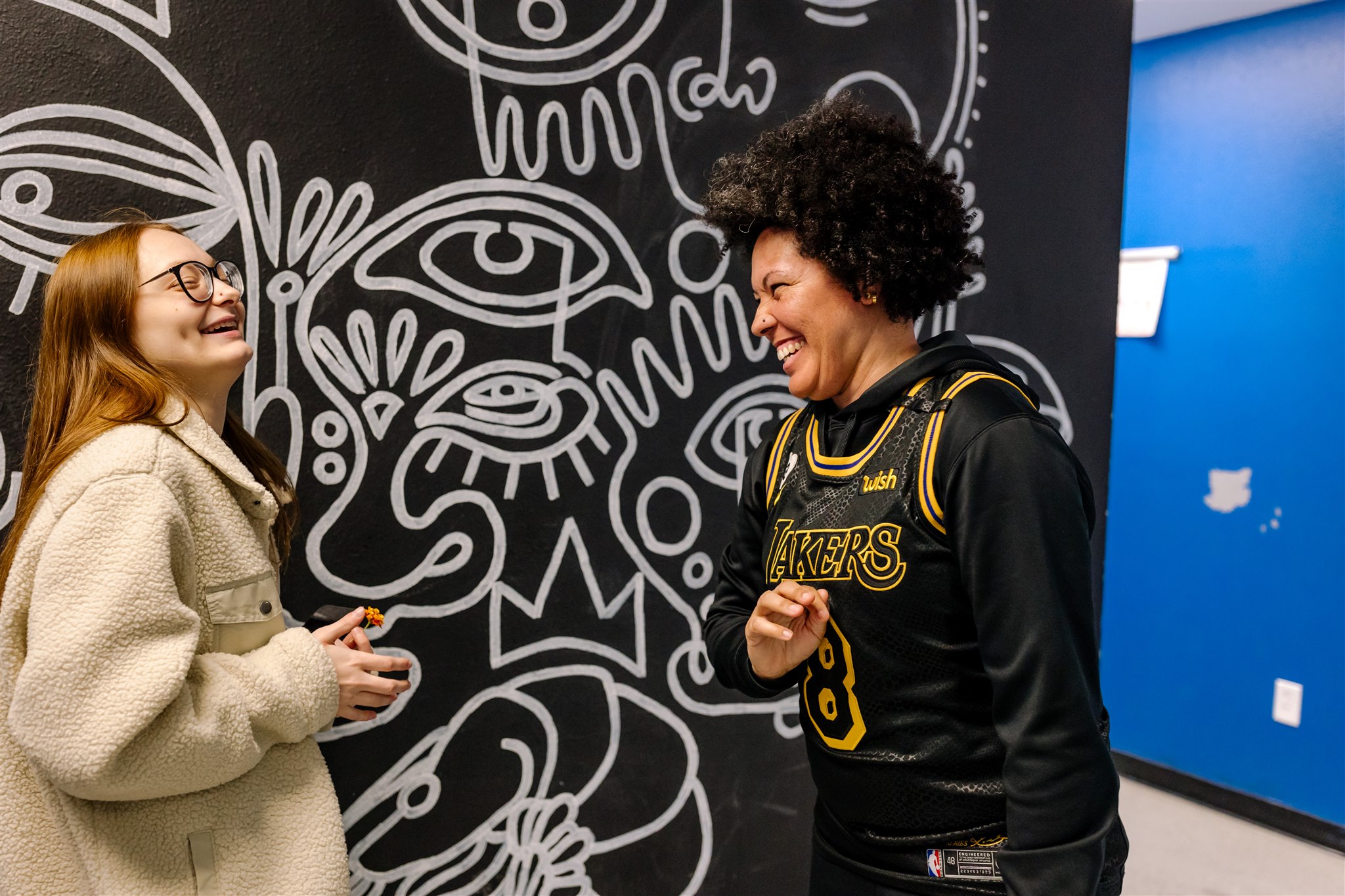 A SET High student laughs with SET High Teacher, Ms. Nicky Geis, in front of a black and white mural.