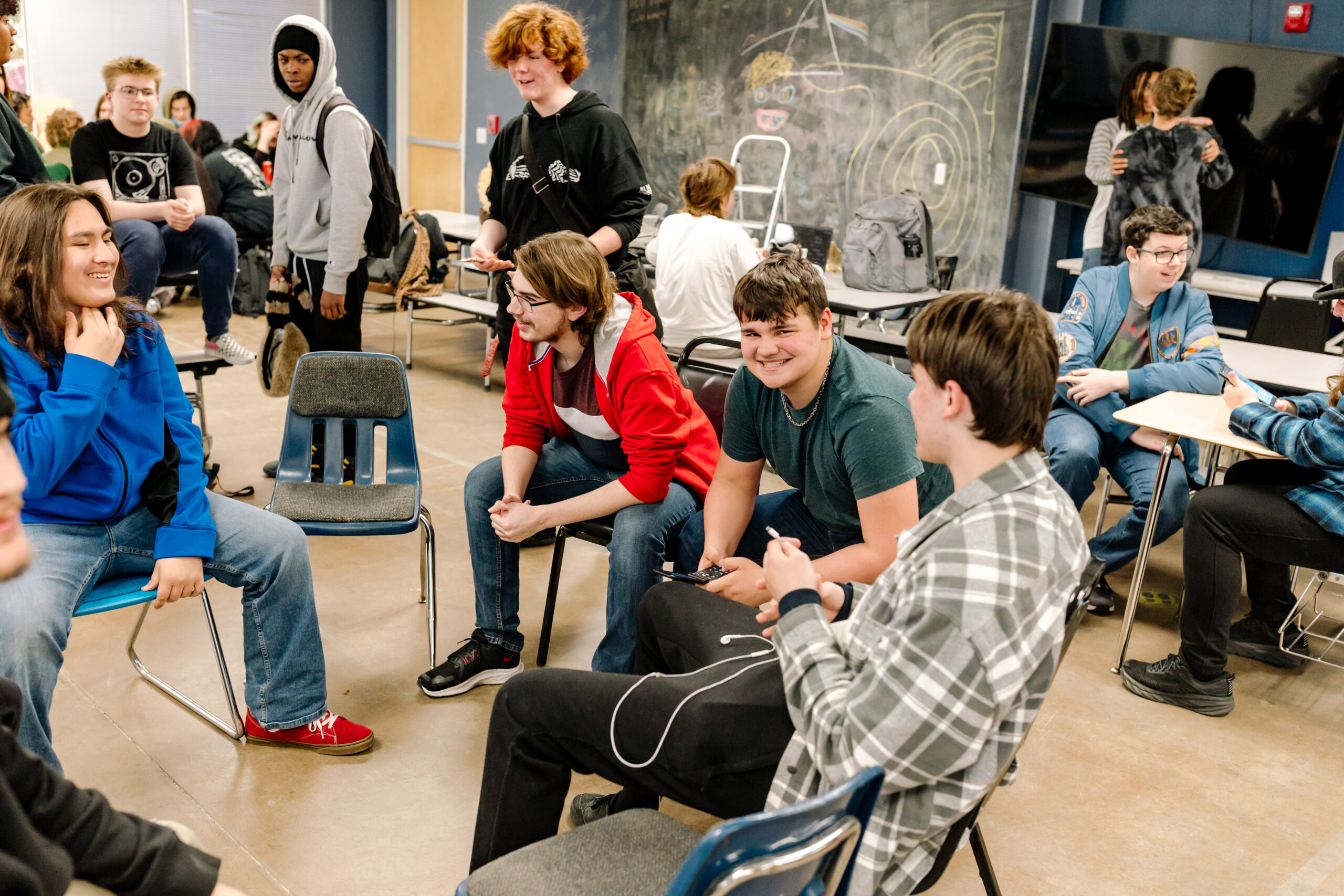 A group of SET High students chat while sitting in chairs in a circle with other students milling around behind them.
