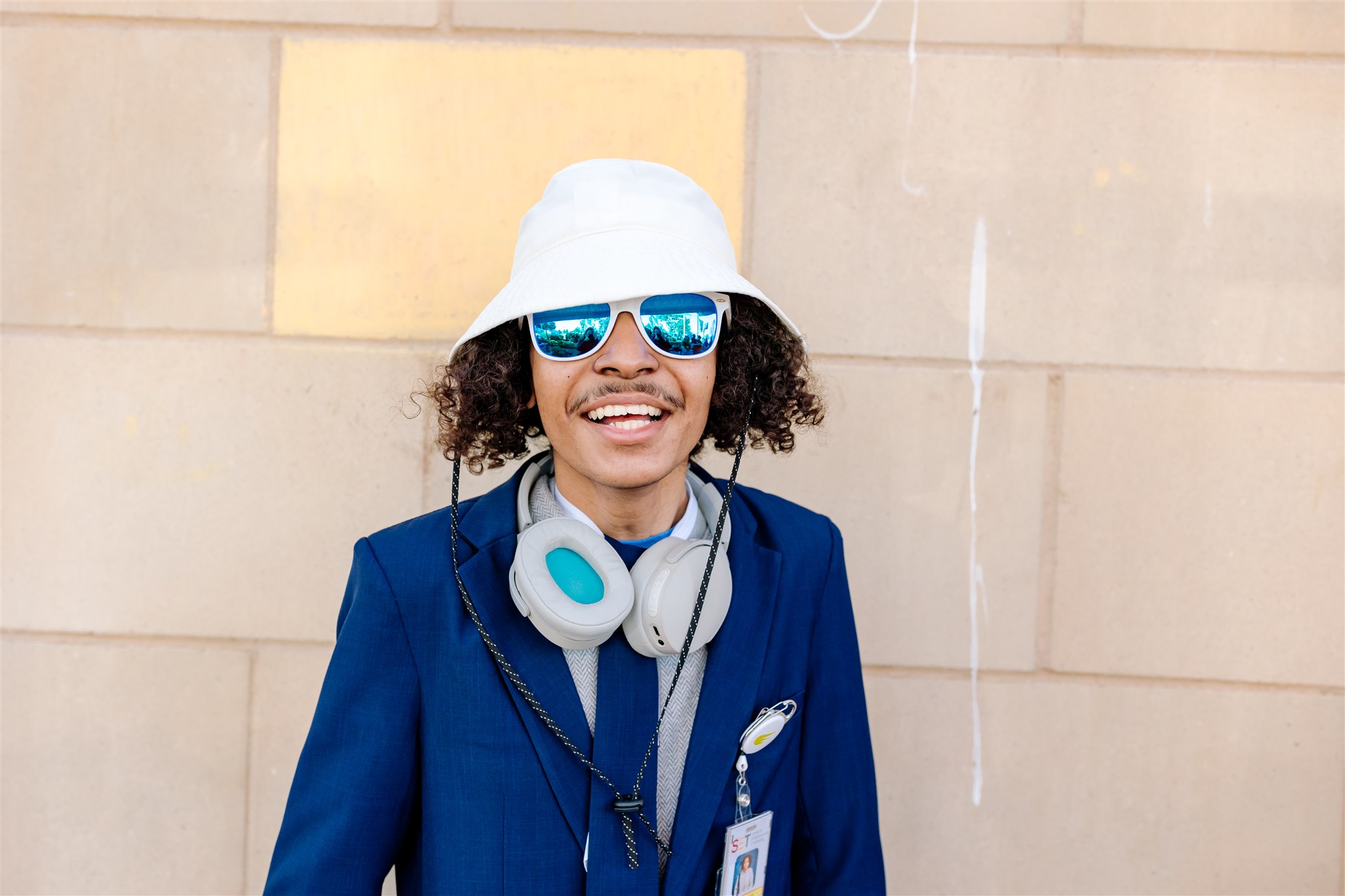 SET High student wearing a bright blue blazer, white bucket hat, mirror blue sunglasses and headphones around their neck smiles for the camera.