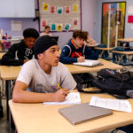 The School for Entrepreneurship & Technology students sit at tables in a classroom and write with pencils on paper while looking off-camera to the left.