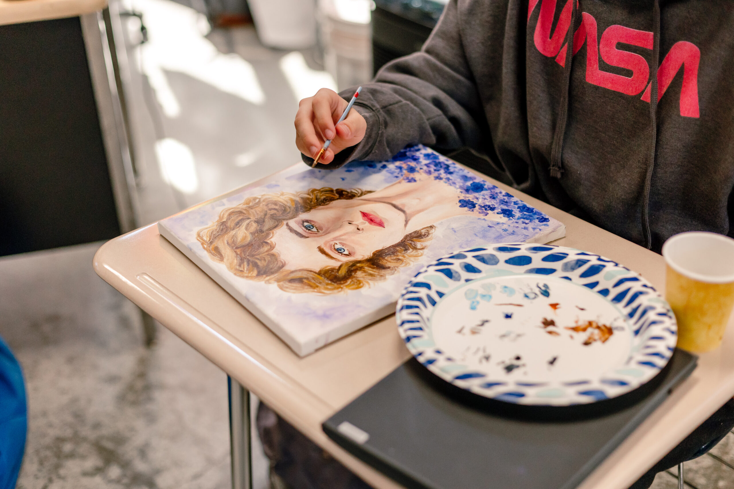 A student at the School for Entrepreneurship and Technology sits at a desk with a paintbrush in hand and a small canvas with a painted face on the desk in front of them.