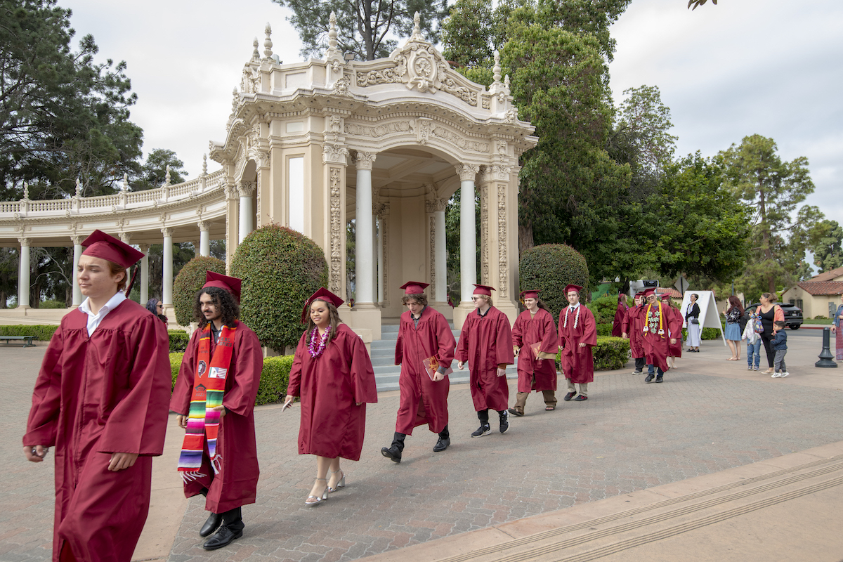 A line of The School for Entrepreneurship and Technology (SET) graduates walk across an outside stone path in red caps and gowns with an ornate stone colonnade behind them. 