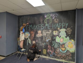 A couple of SET High students draw on an extensive mural chalk wall in multi-colors under a heading that reads, “Art Makes Me Happy”.