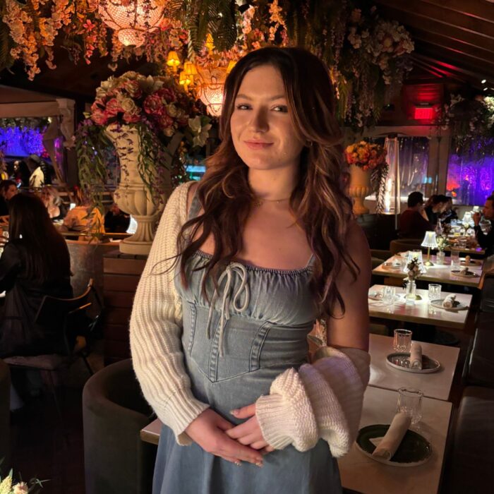 SET High graduate Juniper Davidson stands in a dimly lit restaurant with tables set with white tablecloths and napkins and colorfully lit with lanterns and flowers.
