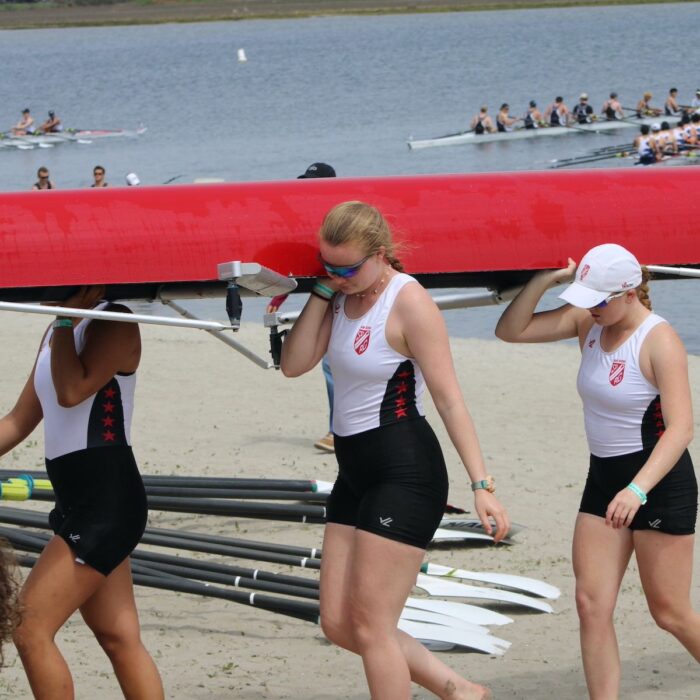 Three rowers (including SET High graduate Mahaila Bouman) carry part of a long red row boat on their shoulders with a lake, boats and rowers in the background.