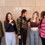 SET High students stand close to one another in front of an outside wall in a line, some looking and smiling at the camera and a couple looking to the side.